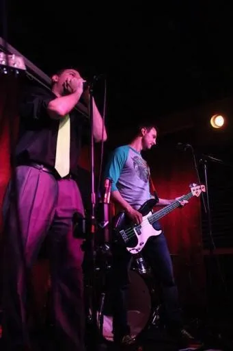 2 musicians performing on a dark stage, 1 singing and 1 playing bass guitar
